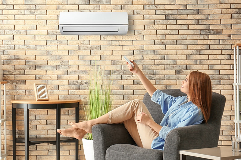 Young woman switching on air conditioner while sitting in armchair at home.