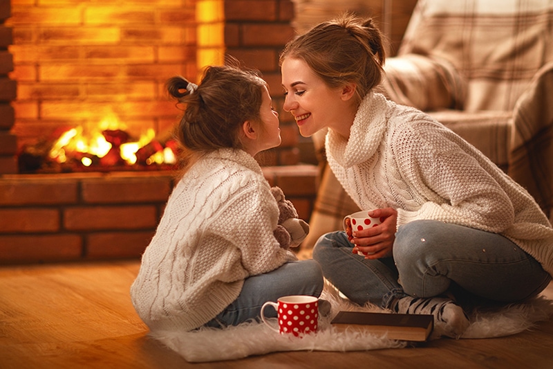 Mother and daughter snuggled up in front of a fireplace with mugs of hot cocoa.